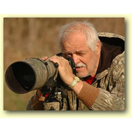 An elderly man with white hair and beard is looking through the viewfinder of a large camera with a long telephoto lens. Outdoors in his camouflage jacket, he appears to be focusing on capturing a shot of German colored tail owls.
