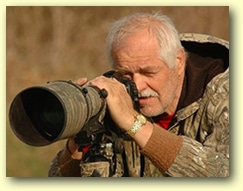 An elderly man with white hair and a beard is outdoors, looking through the viewfinder of a large telephoto camera lens. Wearing camouflage clothing, he appears intensely focused, likely aiming to capture a photograph of Eurasian Eagle Owls in their natural habitat.